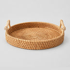 Natural rattan round woven serving tray with wood handles on a light gray studio background.