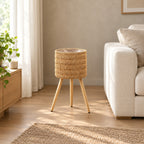 Standing woven rattan planter basket on wooden tripod legs in Natural Rattan on the floor beside a 200 cm wide cream sofa in a calm room.