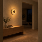 Round travertine disc wall sconce lit in the evening mounted on the plaster wall of a hallway beside an 80 cm wide doorway, the only light in the room