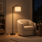 Matte Black arched floor lamp lit in the evening standing on the floor beside an 80 cm wide boucle armchair in a reading corner, the only light in the room.